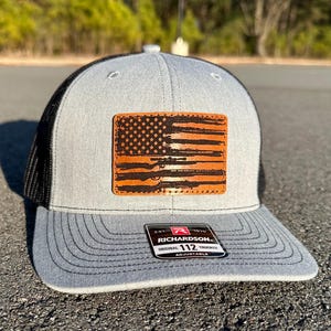 Könnte beinhalten: Eine graue und schwarze Trucker-Kappe mit einem braunen Leder-Patch der amerikanischen Flagge. Der Patch hat einen Used-Look und zeigt die Silhouette eines Gewehrs. Die Kappe ist verstellbar und hat den Text "Richardson 112" auf der Rückseite.