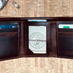 Op de afbeelding: Open bruin leren drievoudige portemonnee met meerdere kaartsleuven en een doorzichtig ID-venster. De portemonnee is opengeklapt, waardoor de binnencompartimenten zichtbaar zijn. Een zilveren horloge en een ring zijn op de achtergrond zichtbaar.