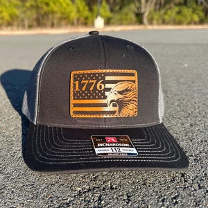 Puede incluir: Gorra de camionero negra y gris con un parche de cuero con el número "1776" y un diseño de cabeza de águila. La gorra tiene una visera curva y una correa ajustable.