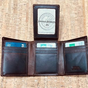 May include: Open brown leather trifold wallet with multiple card slots and a clear ID window. The wallet is open, displaying credit cards and the interior. The text "Emerald Engraving Co." is visible.