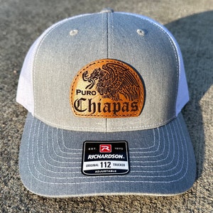 May include: Gray and white trucker hat with a brown leather patch featuring an eagle design and the text "PURO Chiapas". The hat has a black adjustable strap with the text "EST. 1970 RICHARDSON. ORIGINAL 112 TRUCKER ADJUSTABLE".