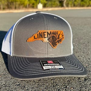 Può includere: Un cappello da camionista grigio e bianco con una toppa in pelle marrone che presenta il contorno della Carolina del Nord e il testo "LINEMAX". Il cappello è regolabile e ha un'etichetta Richardson 112.