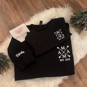 May include: Black long-sleeved shirt and short-sleeved shirt with white text and arrow designs. The long-sleeved shirt has the name "Frieda" on the sleeve. The short-sleeved shirt has the text "MAMA EST 2025" and "MINI EST 2025".