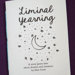 May include: A white zine titled "Liminal Yearning" with a black illustration of a crescent moon and stars. The text "A story game zine about dreams and romance by Eliot Crow" is printed below the illustration. The zine is on a purple surface.