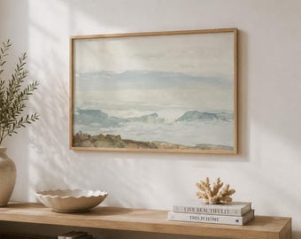 May include: A rectangular landscape painting in a light wood frame featuring muted blue, beige, and brown watercolor tones. The artwork depicts a coastal scene with soft horizon lines, displayed above a long wooden console table with decor.