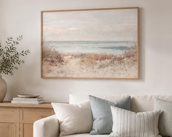 May include: A framed landscape painting of a coastal beach scene with soft beige, blue, and white tones hanging above a white sofa. The artwork depicts sand dunes, tall grasses, and a calm ocean horizon under a muted, cloudy sky in a horizontal orientation.