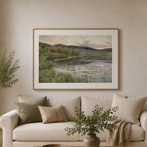 May include: A living room with a cream sofa, cushions, and a landscape painting in a wooden frame. A wooden coffee table holds a bowl and books. A tall plant and a lamp are also visible. The room has a neutral colour palette.