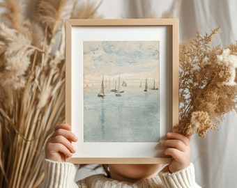 May include: A framed watercolor print of sailboats on the water, with a light wood frame. The print has a soft, muted color palette of blues, greens, and beige. The word "AQUAMARINE" is printed at the top. The frame is held by a person wearing a cream-colored sweater.