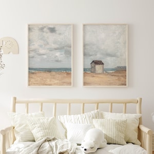 May include: Two framed paintings of a beach scene with a small house on the shore. The paintings are hung on a white wall above a wooden daybed with white and green pillows.