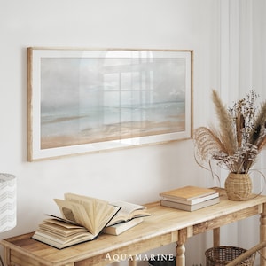 Puede incluir: Una impresión enmarcada de una escena de playa cuelga de una pared blanca. Una consola de madera exhibe libros abiertos, libros apilados y un jarrón tejido con flores secas. La palabra "AQUAMARINE" está impresa en la mesa.