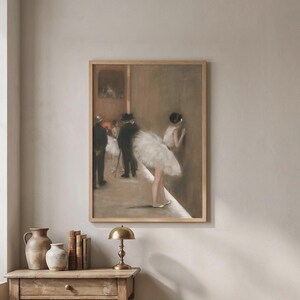 May include: A framed wall art print depicting a ballet scene with dancers in white tutus and figures in dark suits, rendered in muted earth tones. The artwork is displayed in a light wood frame, capturing a soft, painterly aesthetic with light.