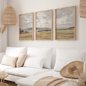 Könnte beinhalten: Drei gerahmte Landschaftsbilder mit einer gedämpften Farbpalette, die ein Feld mit einem Gewässer in der Ferne darstellen. Die Bilder hängen an einer weißen Wand über einem weißen Sofa mit beigen Kissen.