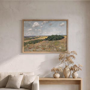 May include: A living room scene featuring a beige sofa with throw pillows, a wooden console table, and a framed landscape painting. The painting depicts a field with trees under a cloudy sky. Dried flowers in vases add a natural touch.