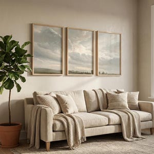 May include: A neutral-toned living room with a beige sofa, cushions, and throws. Three framed landscape prints are mounted on the wall. A potted plant and a rug complete the scene.