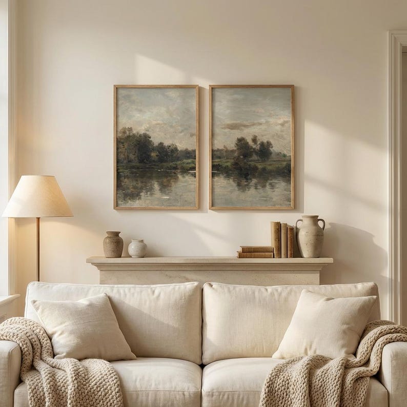 May include: Two framed landscape paintings depict a serene river scene with trees and a cloudy sky. The artwork is displayed above a cream-colored sofa with throw pillows and a knitted blanket. A floor lamp and decorative vases complete the room.