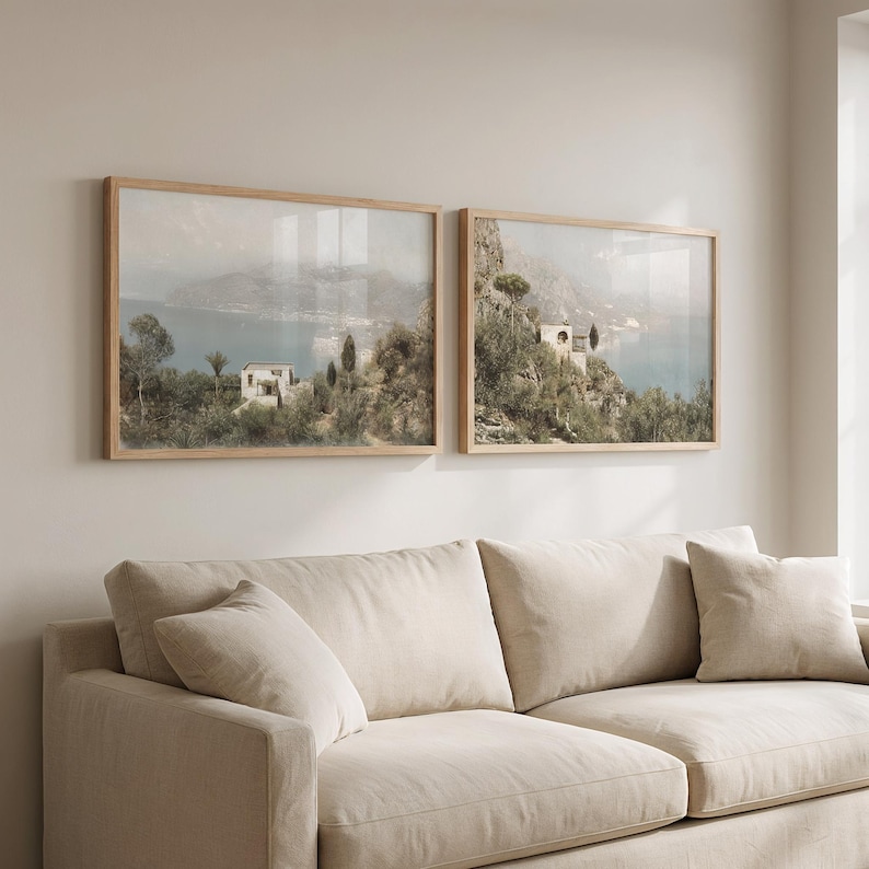 May include: Two framed landscape prints showing coastal scenes with buildings, trees, and water. The prints have light wood frames and are mounted on a neutral wall above a beige sofa with cushions.
