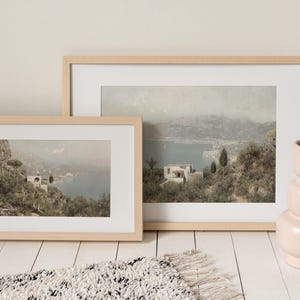 May include: Two framed landscape prints in light wood frames. The prints show a coastal scene with buildings, trees, and water. A pink vase with dried branches is on the right. A white and black rug is in the foreground.