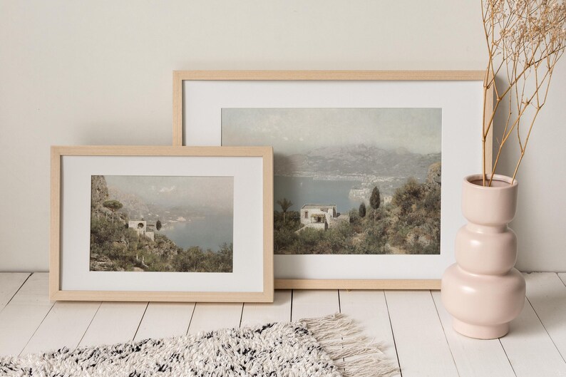 May include: Two framed landscape prints in light wood frames. The prints show a coastal scene with buildings, trees, and water. A pink vase with dried branches is on the right. A white and black rug is in the foreground.