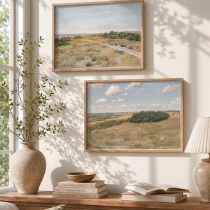 May include: Two framed landscape paintings with wooden frames hang on a cream-colored wall. The paintings depict fields and a dirt road under a blue sky. A vase with greenery, books, and a lamp sit on a wooden table.