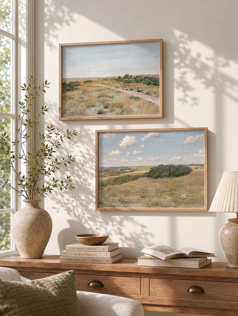 May include: Two framed landscape paintings with wooden frames hang on a cream-colored wall. The paintings depict fields and a dirt road under a blue sky. A vase with greenery, books, and a lamp sit on a wooden table.