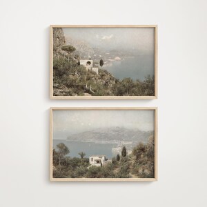 May include: Two framed landscape prints. Each print shows a coastal scene with buildings among greenery. The artwork uses muted green, blue, and beige tones, giving a vintage feel. The frames are made of light-coloured wood.
