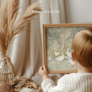 May include: A framed watercolor print featuring a pastoral scene with white geese. The artwork is held by a person, with the text "AQUAMARINE" at the top. The frame is a light wood color, and the artwork has a vintage aesthetic.