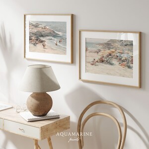May include: Two framed beach scene prints with light wood frames hang on a white wall. The prints depict people on a sandy beach with ocean waves and colorful umbrellas. A beige lamp and desk are in the foreground.