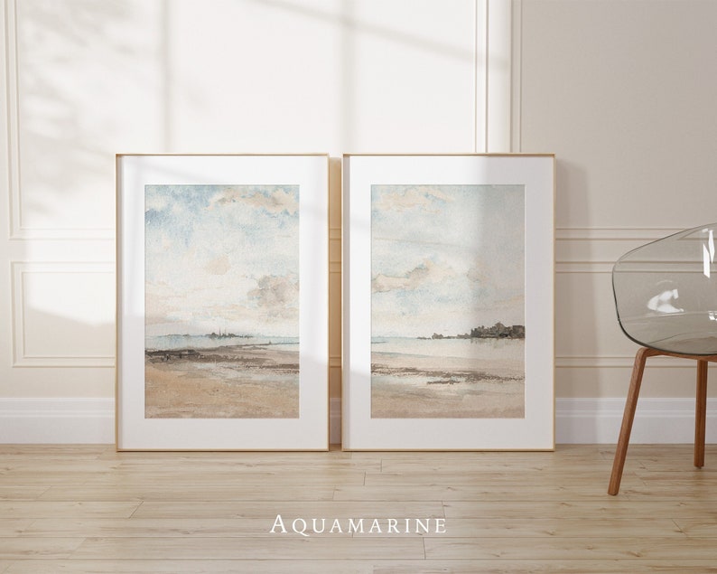 May include: Two framed watercolor paintings of a seascape with a light blue sky, white clouds, and a sandy beach. The paintings are in gold frames and are hanging on a white wall.