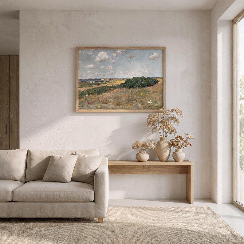 May include: A living room scene featuring a beige sofa with throw pillows, a wooden console table, and a framed landscape painting. The painting depicts a field with trees under a cloudy sky. Dried flowers in vases add a natural touch.