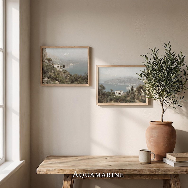 May include: Two framed landscape prints depicting a coastal scene with a light blue sea, white buildings, and green foliage. The prints are in light brown wooden frames. The word "AQUAMARINE" is printed on the wooden table.