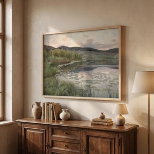 May include: A framed landscape painting shows a tranquil lake scene with lily pads, verdant foliage, and distant hills beneath a soft, cloudy sky. The artwork is displayed above a wooden cabinet with decorative vases and books.