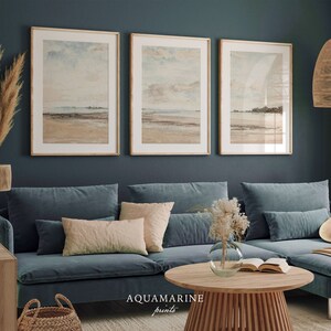 May include: Three framed landscape prints with a beach scene, displayed above a blue velvet sofa. The prints have a neutral color palette with beige, blue, and brown tones. The text "AQUAMARINE prints" is visible at the bottom. A round wooden coffee table and decorative items complete the room.