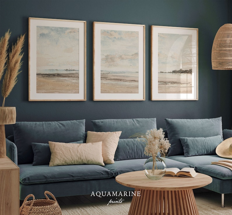 May include: Three framed landscape prints with a beach scene, displayed above a blue velvet sofa. The prints have a neutral color palette with beige, blue, and brown tones. The text "AQUAMARINE prints" is visible at the bottom. A round wooden coffee table and decorative items complete the room.