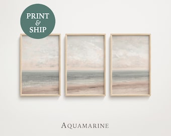 May include: Three vertical framed wall art prints featuring a continuous coastal landscape with a soft pink sky, muted blue ocean waves, and a sandy beach. The word AQUAMARINE is printed in black below the frames, and a circular badge says PRINT & SHIP.