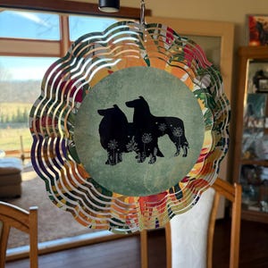 May include: A decorative wind spinner featuring a layered, wavy design with a central circular image. The image showcases black silhouettes of two dogs on a green background with floral accents. The outer edges display a colorful floral pattern.