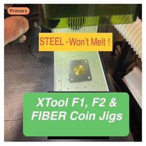 May include: A square steel coin jig with a circular center cutout, designed for XTool F1, F2, and fiber laser machines. The metal plate features four corner holes and is positioned under a green laser shield. Text reads: STEEL - Won't Melt! XTool F1, F2 & FIBER Coin Jigs.