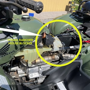 May include: Close-up of a green Honda Rancher ATV with a white arrow pointing to a mouse blocker on the gas tank. The mouse blocker is a small, black plastic device that is designed to prevent mice from entering the engine compartment.