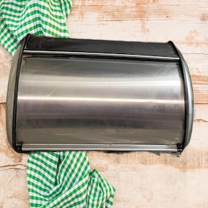 May include: A stainless steel bread box with a black top and curved edges. A green and white checkered cloth is draped on a wooden surface near the bread box. The bread box has a shiny, reflective surface.