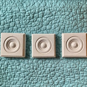 May include: Three square, white wooden decorative blocks with a circular, raised design in the center. The blocks are arranged in a row and sit on a textured, light blue surface. The blocks have a slightly distressed finish.