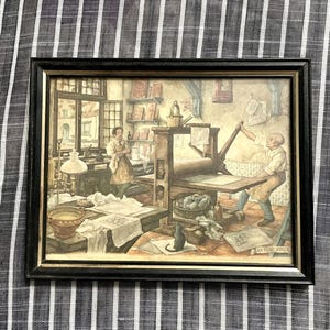 May include: Framed print depicting a vintage print shop scene. The artwork features two figures working with a printing press, surrounded by books, papers, and tools. The print is in a black frame.