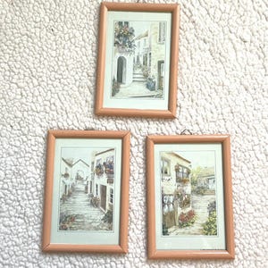 May include: Three framed prints featuring watercolor paintings of European village scenes. Each print is in a light brown wooden frame and depicts a cobblestone street with buildings, flowers, and architectural details. The prints are displayed on a white, textured surface.
