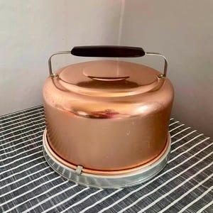May include: A round copper cake carrier with a smooth, metallic finish and a flat top. It features a dark brown rectangular handle attached to a silver-toned metal frame. The carrier sits on a circular silver-toned base with a simple locking mechanism.