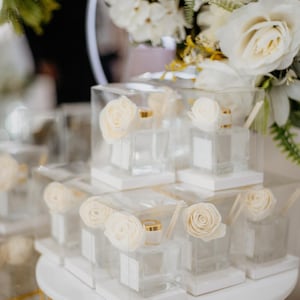Peut inclure: Flacons diffuseurs en verre transparent avec des couvercles dorés et des bâtonnets en bois, présentés dans des boîtes transparentes. Chaque boîte présente une rose crème. Les flacons sont disposés sur un présentoir blanc à plusieurs niveaux avec une base dorée.