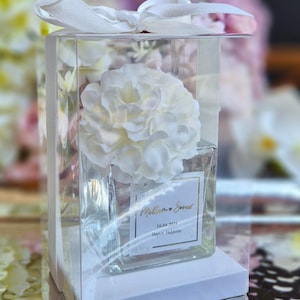 May include: A clear glass jar with a white flower inside. The jar has a white label with the text "William & Sarah 10.04.2023  Nigel Hallam" in gold lettering.