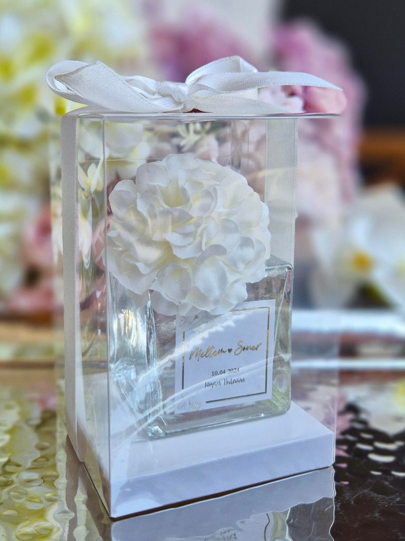 May include: A clear glass jar with a white flower inside. The jar has a white label with the text "William & Sarah 10.04.2023  Nigel Hallam" in gold lettering.