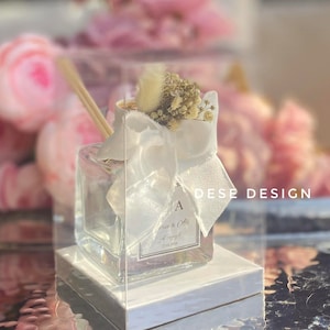 May include: A clear glass bottle diffuser with a white ribbon and dried flowers. The bottle has a label with the text "Jane & Ollie" and the date "27.01.2024". The bottle is sitting on a white square base.