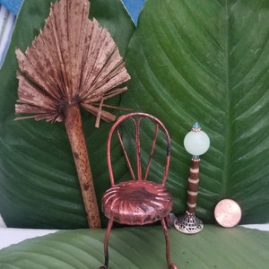 May include: A miniature metal chair with a copper finish sits on a green leaf. A small lamp with a white glass shade and a copper base is next to the chair. A brown palm frond is in the background.