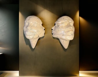 May include: Two white, textured face sculptures mounted on a dark wall. Each sculpture features two faces merged together, with a pointed base. The sculptures are lit by spotlights, creating shadows and highlighting the details.