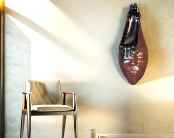 Könnte beinhalten: Moderne Innenraumgestaltung mit einer Wandskulptur eines Gesichts und einer Hand in Braun- und Schwarztönen. Ein Holzstuhl mit cremefarbenem Kissen und zwei kleine dekorative Gegenstände befinden sich im Vordergrund.