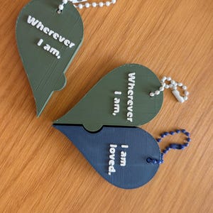 Peut inclure: Trois porte-clés en forme de cœur avec des chaînes à billes blanches. Un cœur vert olive porte l'inscription "Wherever I am,". Un autre cœur vert olive porte "Wherever I am," et un cœur bleu foncé porte "I am loved.", formant un puzzle.